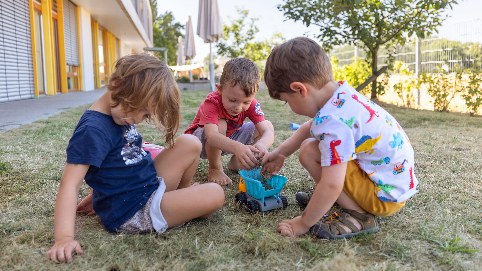 Die Kinder der SGA Sport-Kita spielen zusammen draußen im Garten mit einem Spielzeug-Lastwagen.